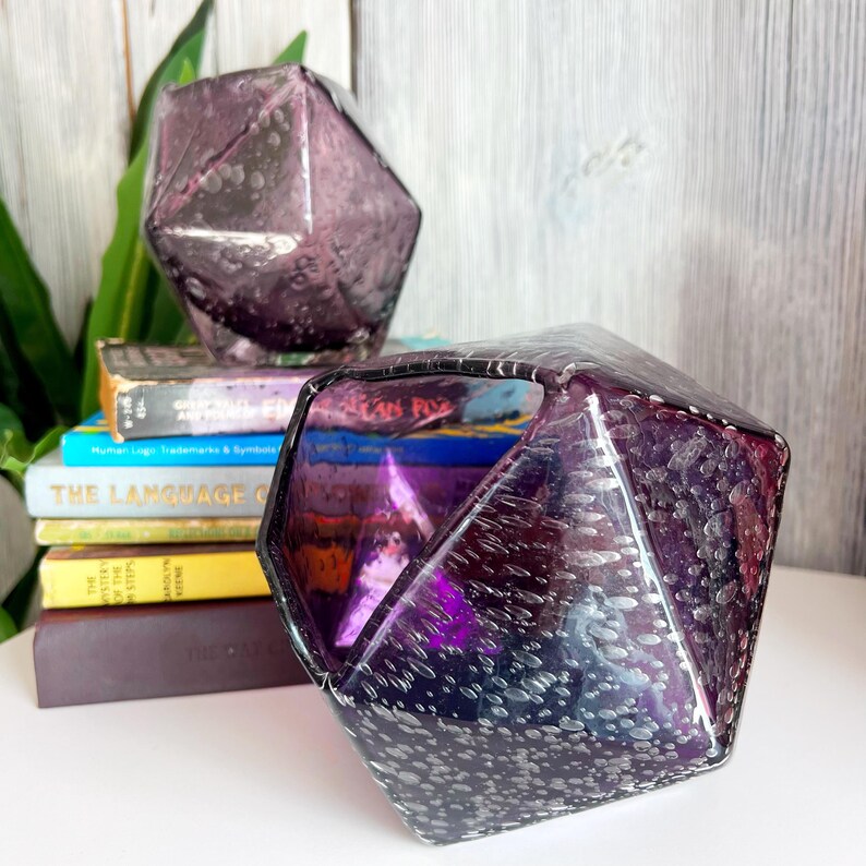 Puede incluir: Dos objetos decorativos de vidrio facetado de color amatista con un patr&oacute;n moteado. Uno se encuentra encima de una pila de libros, el otro en primer plano. Los libros tienen cubiertas coloridas y t&iacute;tulos como "The Language Of".