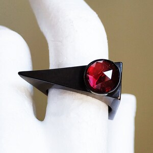 May include: A black ring with a red gemstone set in a triangular shape.