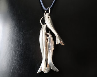 Könnte beinhalten: Silberne Halskette mit einem Anhänger aus drei silbernen Fischen. Die Fische sind übereinander gestapelt, wobei der Schwanz des oberen Fisches nach oben und der Schwanz des unteren Fisches nach unten zeigt. Die Halskette befindet sich auf einem schwarzen Hintergrund.
