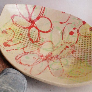 May include: A ceramic bowl with a leaf-shaped design. The bowl is decorated with a red and yellow floral pattern on a cream background.