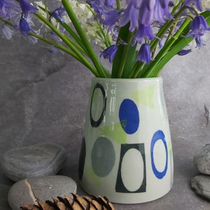 May include: A white ceramic vase with a geometric pattern of blue, green, and gray circles and squares. The vase is filled with a bouquet of purple bluebells.