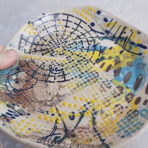May include: A white ceramic plate with a unique abstract design featuring yellow, blue, and brown paint splatters and geometric patterns.