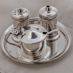May include: A silver condiment set on a round tray. The set includes two shakers and a small lidded container with a spoon. The items are highly reflective, with a bright, polished finish. The tray and containers are made of metal.