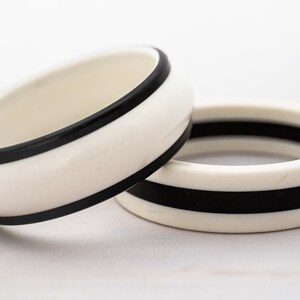 May include: Two white bangle bracelets with black stripes. The bracelets are made of a smooth, glossy material.