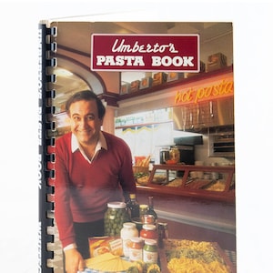 May include: The cover of Umberto's Pasta Book shows a man in a red jumper standing in a restaurant kitchen. The book title is in red and white lettering. The sign above the counter reads "hot pasta".