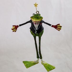 May include: A hanging ornament shaped like a green frog wearing a black tuxedo jacket, a white shirt with a pink bow tie, and green flippers on its feet. The frog has its arms outstretched and a small gold star on a wire above its head.