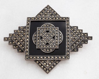 May include: A vintage silver-toned brooch with a geometric design. The brooch features a central black square with a smaller square and circle design, surrounded by marcasite stones. The outer frame has a stepped, Art Deco style.