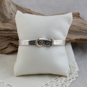 Peut inclure: Un bracelet en argent avec un fermoir en forme de boucle. Le bracelet est sur un coussin blanc.