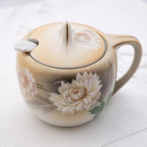 Peut inclure: Un sucrier en porcelaine blanche avec un couvercle et une anse. Le bol est décoré d'un motif floral de chrysanthèmes blancs sur fond brun. Le couvercle est orné d'une bordure dorée et d'une petite cuillère blanche.