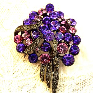 May include: A vintage brooch with a cluster of purple and pink rhinestones. The brooch has a dark metal base with ornate detailing and a flowing, leaf-like design. The rhinestones vary in size and shade, creating a sparkling effect.