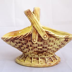 May include: A ceramic basket-shaped planter with a woven texture in shades of yellow and brown. The basket has a handle and a pedestal base. The interior is a lighter yellow.