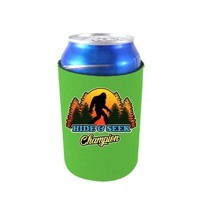 May include: Green can cooler with a silhouette of Bigfoot walking in front of a sunset with trees on either side. The text "HIDE & SEEK Champion" is printed below the image.