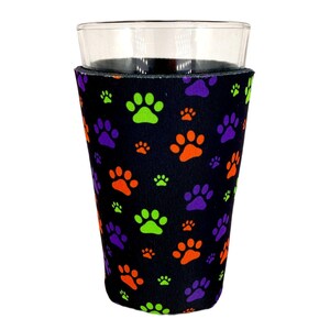 May include: A black drink sleeve with a pattern of orange, green, and purple paw prints. The sleeve is wrapped around a clear glass. The paw prints are in various sizes.