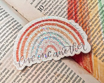 May include: A rainbow-colored sticker with glitter accents. The sticker features a white background with the text "Love one another" in black cursive lettering.