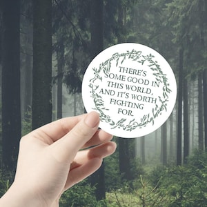 Può includere: Un adesivo rotondo bianco con testo verde e un bordo decorativo. Il testo recita: "C'È DEL BENE IN QUESTO MONDO, E VALE LA PENA COMBATTERE PER ESSO". L'adesivo è tenuto davanti a uno sfondo sfocato di foresta.