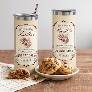 May include: Two cream-colored insulated tumblers with lids and a straw, featuring a label with the text "Green Gables Marilla's Raspberry Cordial." A plate of cookies sits in front of the tumblers. The tumblers are on a wooden surface.