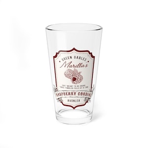 May include: A clear glass pint with a decorative label. The label is maroon and features the text "Green Gables, Marilla's Raspberry Cordial, Avonlea" with a raspberry illustration. The glass is designed for drinking.