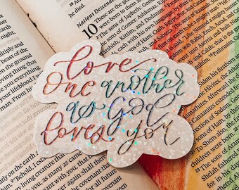 May include: A sparkly, iridescent sticker with a cloud-like shape. The sticker features the text "Love one another as God loves you".