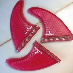 May include: Three translucent pink surfboard fins featuring a repeating hexagonal honeycomb pattern. Each fin displays the white text North East with a small graphic of a person carrying a surfboard. The base of each fin has four cutouts.