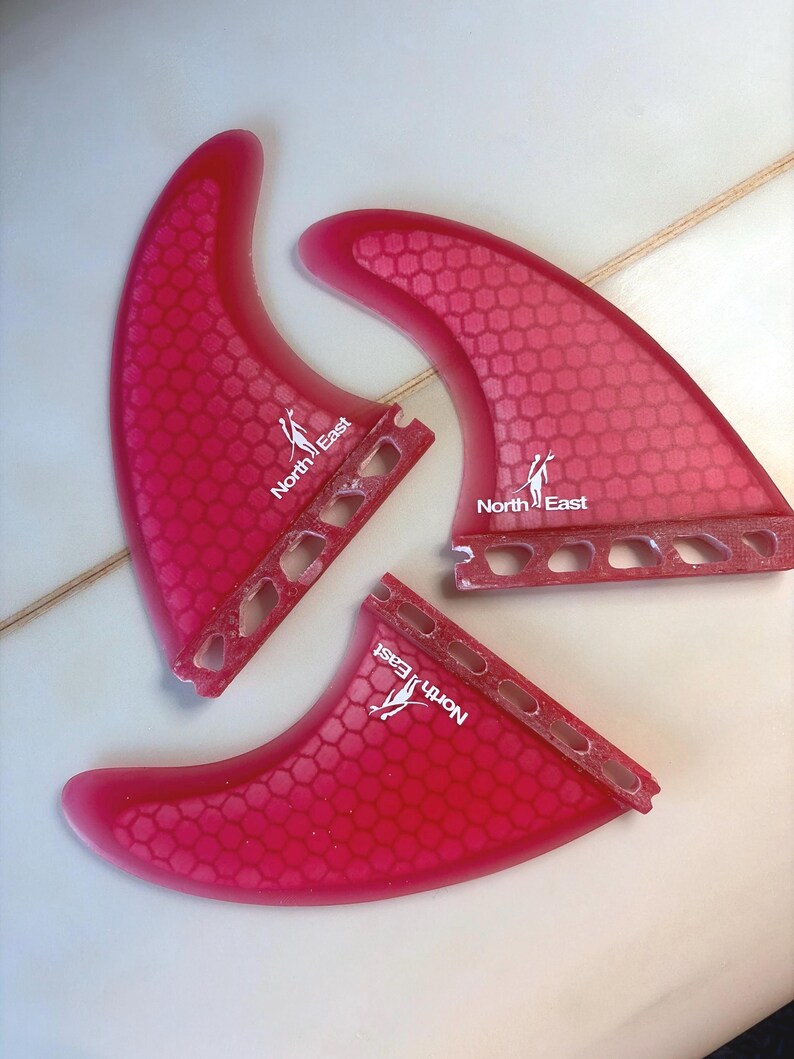 May include: Three translucent pink surfboard fins featuring a repeating hexagonal honeycomb pattern. Each fin displays the white text North East with a small graphic of a person carrying a surfboard. The base of each fin has four cutouts.