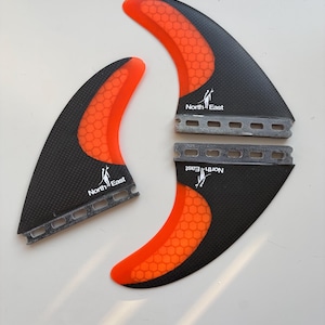 May include: Three surfboard fins featuring a black carbon fiber texture and bright orange honeycomb patterns. Each fin displays the North East logo with a stylized surfer graphic. The base of each fin has a series of rectangular cutouts.
