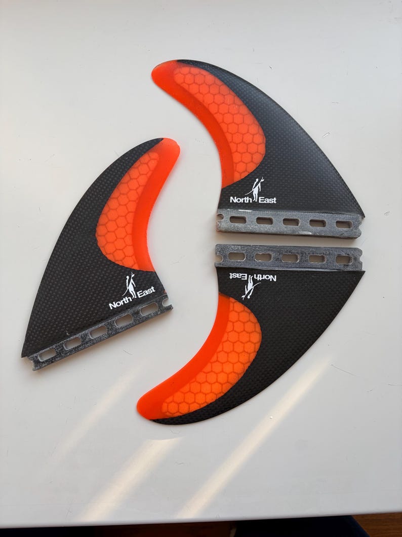 May include: Three surfboard fins featuring a black carbon fiber texture and bright orange honeycomb patterns. Each fin displays the North East logo with a stylized surfer graphic. The base of each fin has a series of rectangular cutouts.