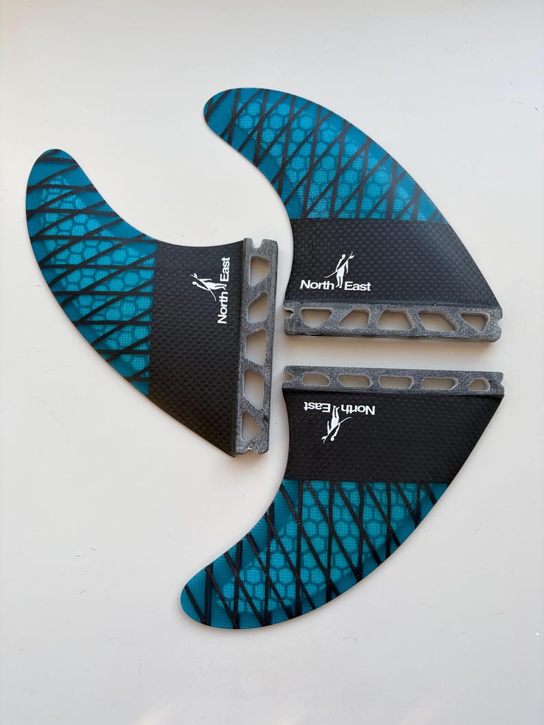 May include: A set of three surfboard fins featuring a teal and black honeycomb pattern. Each fin has a black base with the text North East and a logo of a person surfing. The base of each fin includes a series of cut-out rectangular holes.
