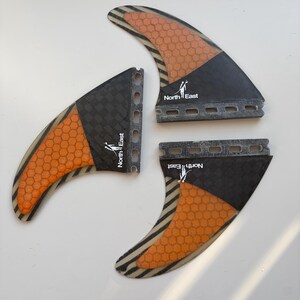 May include: A set of three surfboard fins featuring a black and orange honeycomb pattern. Each fin displays the text North East with a small graphic icon. The fins have a dark carbon fiber texture near the base and a series of mounting holes.