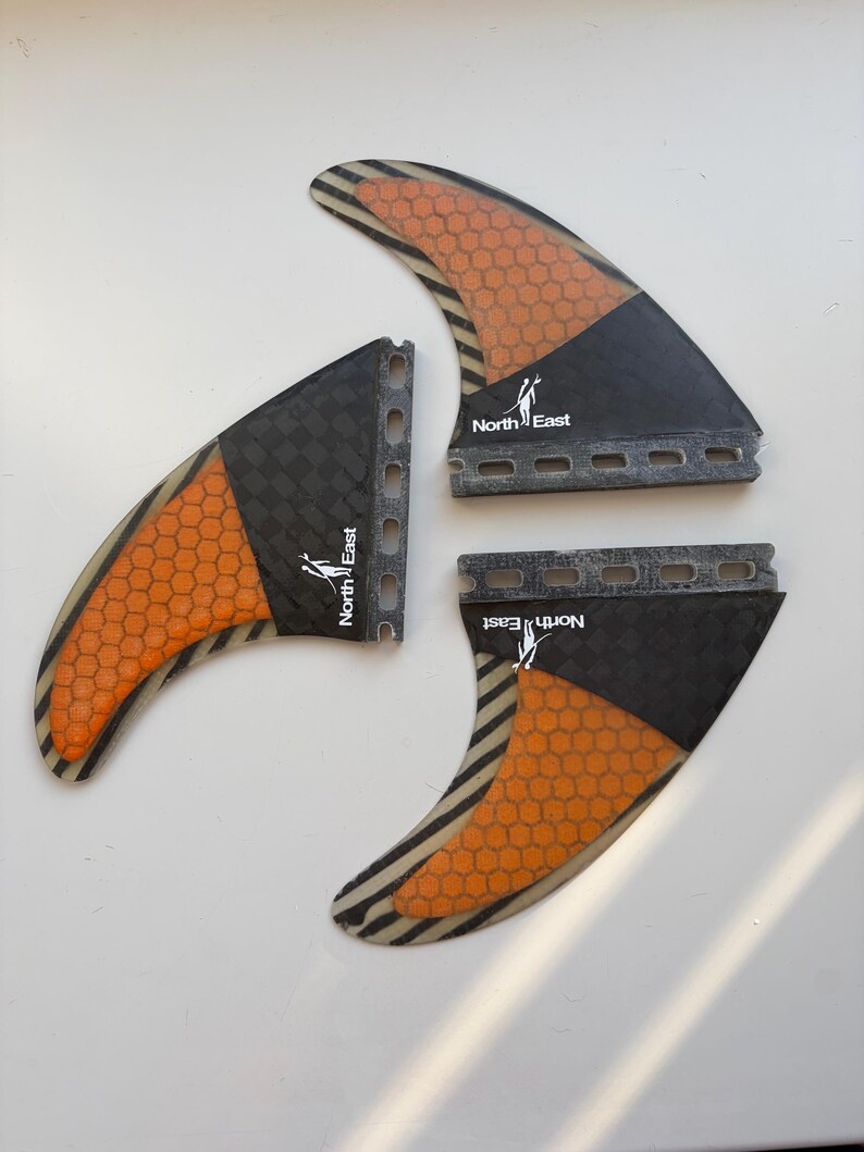 May include: A set of three surfboard fins featuring a black and orange honeycomb pattern. Each fin displays the text North East with a small graphic icon. The fins have a dark carbon fiber texture near the base and a series of mounting holes.