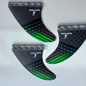 May include: A set of three surfboard fins featuring a black and grey checkered pattern with bright green diagonal stripes. Each fin has a grey base with cutouts and the white text North East with a small logo printed on the surface.