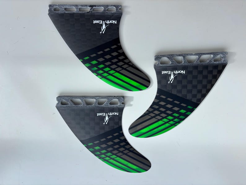 May include: A set of three surfboard fins featuring a black and grey checkered pattern with bright green diagonal stripes. Each fin has a grey base with cutouts and the white text North East with a small logo printed on the surface.