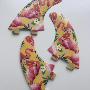 Op de afbeelding: Drie surfvinnen met een flamingopatroon. De vinnen tonen roze flamingo's, groene bladeren en witte bloemen op een gele achtergrond. De vinnen zijn ontworpen voor surfboards.