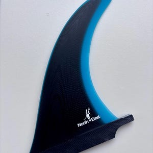 May include: A surfboard fin featuring a black textured base that transitions into a translucent blue tip. The fin displays a white logo with the text North East and a small graphic of a person on a surfboard near the bottom edge of the black section.