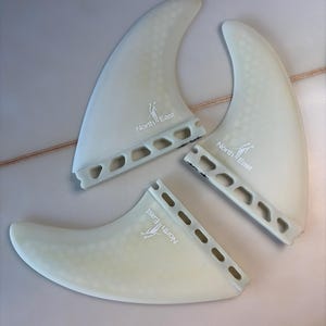 May include: A set of three translucent off-white surfboard fins featuring a honeycomb pattern and the North East logo printed in white. Each fin has a base with a series of rectangular cutouts along the attachment edge for mounting to a board.