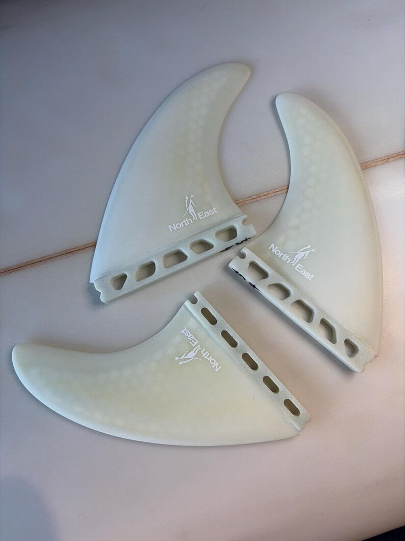 May include: A set of three translucent off-white surfboard fins featuring a honeycomb pattern and the North East logo printed in white. Each fin has a base with a series of rectangular cutouts along the attachment edge for mounting to a board.