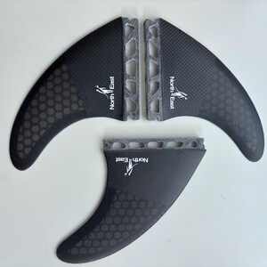 May include: A set of three black surfboard fins featuring a carbon fiber weave pattern, a honeycomb texture on the lower section, and a grey textured base. Each fin displays the white text North East with a small graphic of a person surfing.