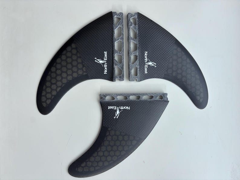 May include: A set of three black surfboard fins featuring a carbon fiber weave pattern, a honeycomb texture on the lower section, and a grey textured base. Each fin displays the white text North East with a small graphic of a person surfing.