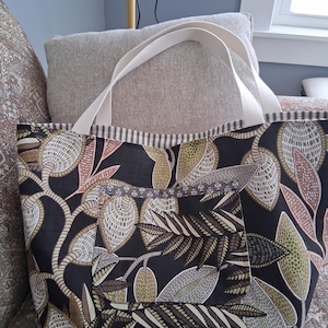 May include: A black tote bag with a botanical print of leaves and branches in brown, beige, and green. It has a cream-coloured handle and a striped border. A pocket is visible on the front.
