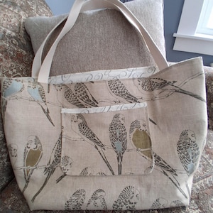Puede incluir: Bolso tote beige con estampado de pájaros. El bolso presenta un patrón repetido de pájaros posados en ramas en tonos marrón, gris y azul. Tiene un bolsillo frontal y asas color crema.