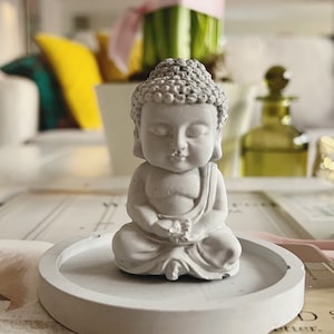 May include: A small, grey Buddha statue in a seated meditation pose. The statue sits on a round, light grey tray. The background includes a vase of purple flowers and a decorative glass bottle.