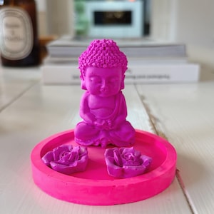 Puede incluir: Una figura de Buda de color rosa neón brillante sentada en posición de meditación sobre una bandeja circular a juego. Dos pequeñas flores de rosa esculpidas en color rosa neón están colocadas sobre la bandeja frente a la figura, creando un conjunto monocromático.