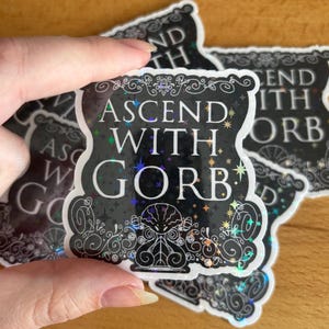 May include: A stack of black and holographic stickers with the text "ASCEND WITH GORB" in white and colorful lettering. The stickers have a decorative border and a stylized illustration.