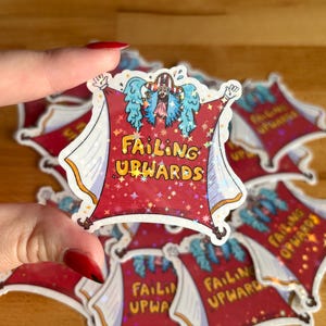 May include: A red sticker with the text "Failing Upwards" in yellow and orange. The sticker features a cartoon character with blue wings and a red cape. The background is a sparkly red with white and gold accents. Other stickers are visible in the background.