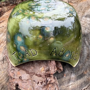 May include: A handcrafted ceramic toad abode with a glossy green glaze. The surface features a mottled pattern of teal and gold. The words "TOAD ABODE" are inscribed on the front. This unique piece is designed for garden decor.