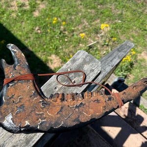 May include: A weathered, dark-colored jawbone with visible teeth, bound with a brown leather strap. The jawbone has a distressed, aged appearance, with areas of rust and discoloration. The object is likely a decorative or artistic piece.