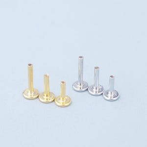 May include: A collection of six flat-back labret studs arranged in two groups. Three studs are gold-toned and three are silver-toned, each featuring a circular base and a vertical post of varying heights, designed for body piercing jewelry.