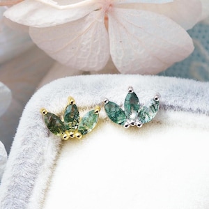 May include: Two stud earrings shaped like small crowns or leaves, each featuring three marquise-cut moss agate stones. One earring is set in yellow gold-toned metal, while the other is set in silver-toned metal, both displayed on a soft gray velvet surface.