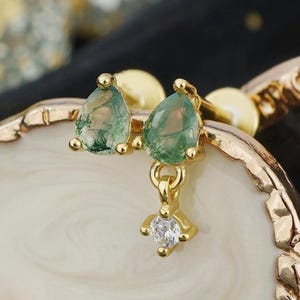 May include: A gold-toned earring featuring two teardrop-shaped green moss agate stones and a small dangling clear cubic zirconia crystal. The earring has a post-back closure and is constructed with a polished metal setting.