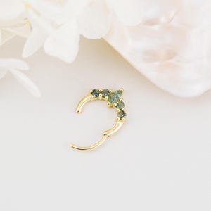 May include: A gold-colored hinged hoop earring with a row of small, green gemstones. The earring is open, showing the clasp mechanism. The background is a soft, neutral color, with a hint of white flowers and a shell-like shape.