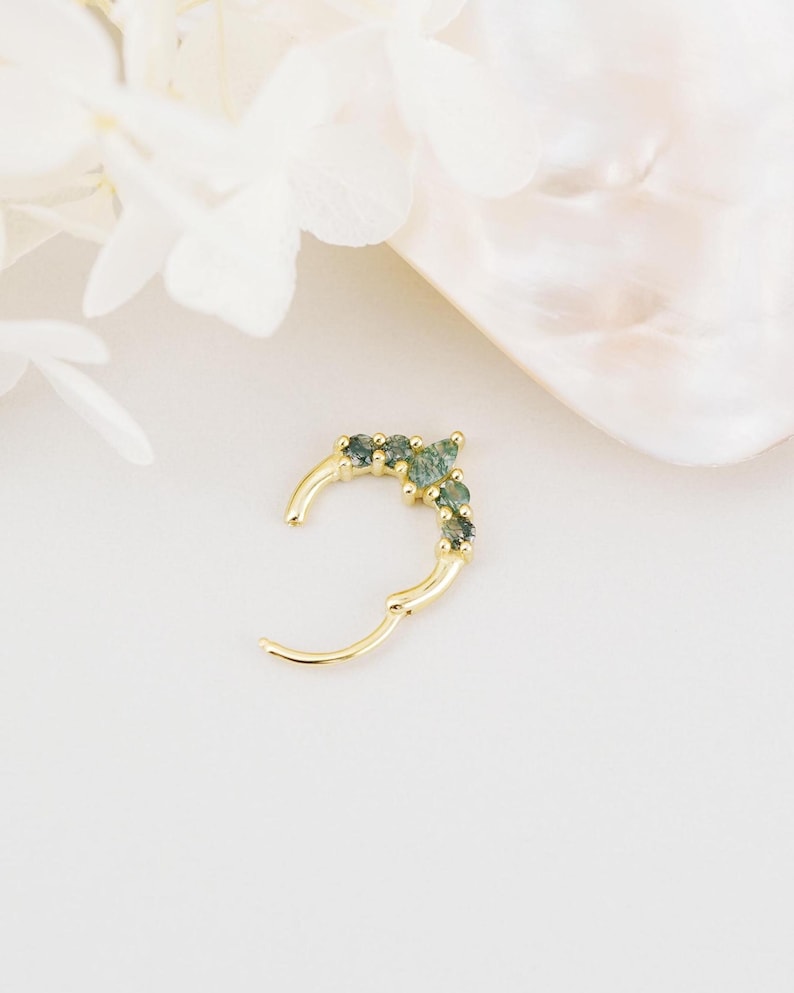 May include: A gold-colored hinged hoop earring with a row of small, green gemstones. The earring is open, showing the clasp mechanism. The background is a soft, neutral color, with a hint of white flowers and a shell-like shape.